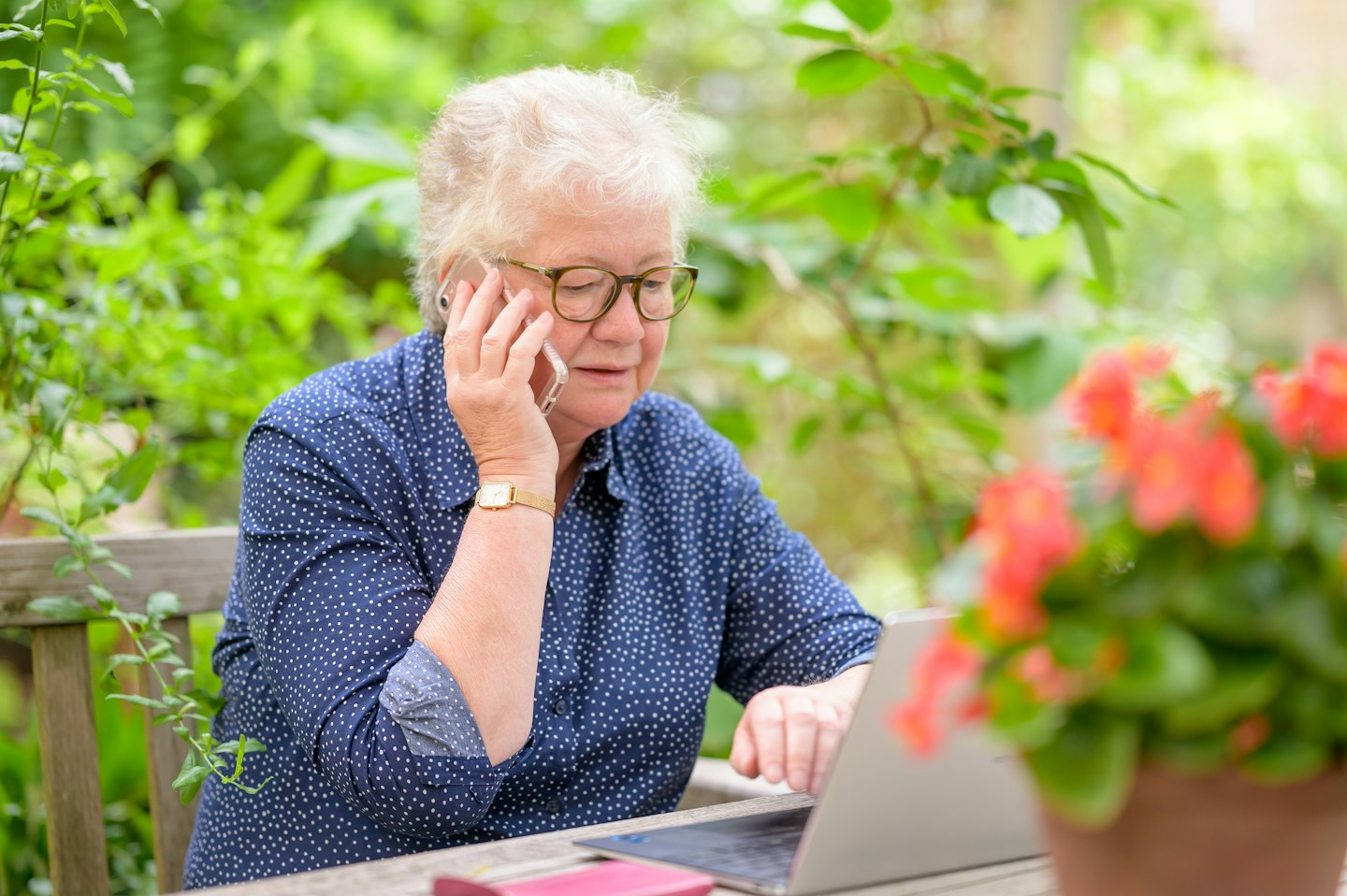 An older woman uses a phone while working at a laptop.